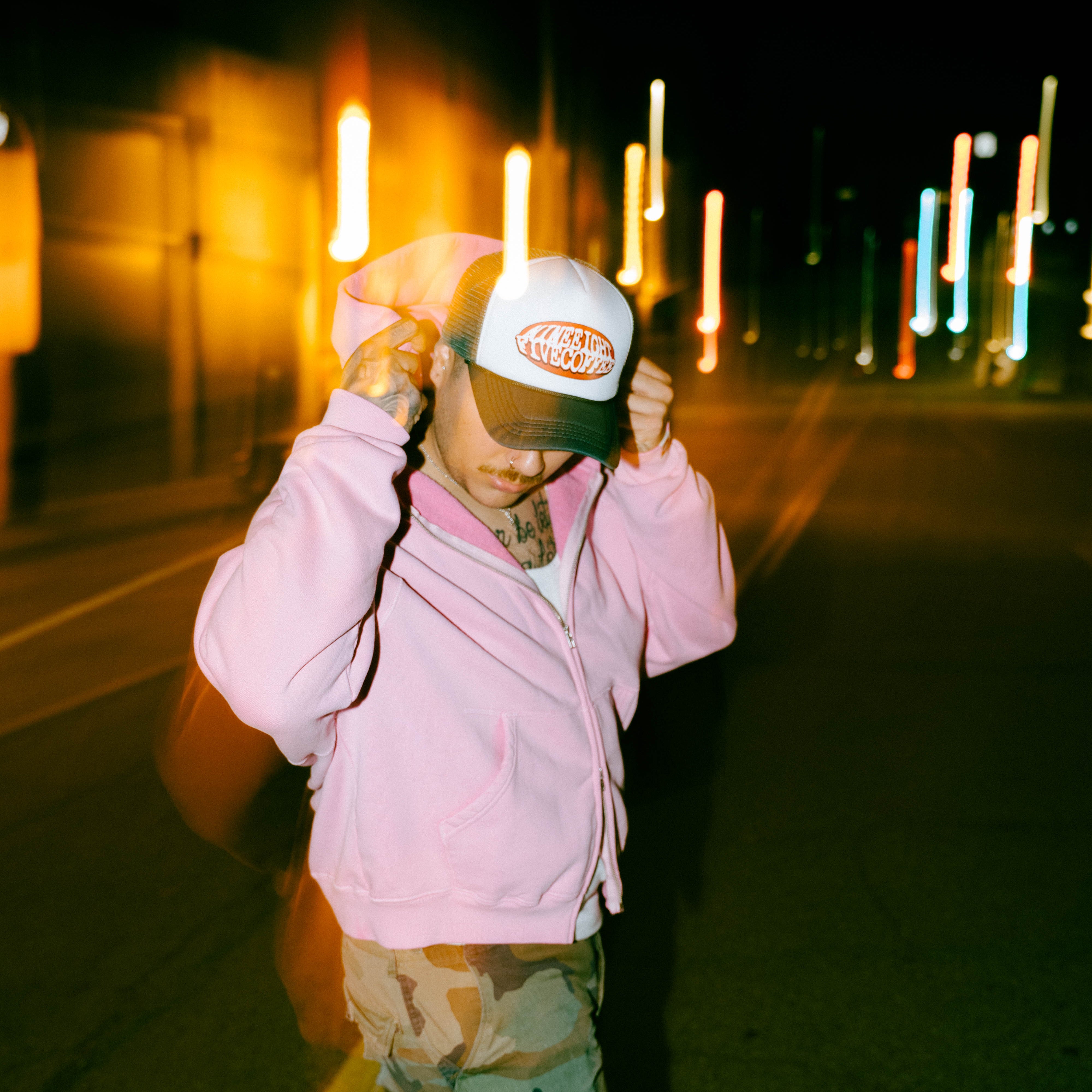 Person wearing a pink jacket and camouflage pants on a dark street with lights in the background